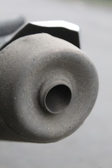 A close-up view of a dusty and slightly weathered motorcycle or vehicle exhaust pipe. The texture is rough with visible specks of dirt, and the pipe's circular opening is prominently featured.