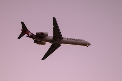 A commercial airplane, branded with Hawaiian Airline's logo and colors, is in mid-flight against a solid purple sky backdrop. The plane is well-illuminated with details visible on its fuselage and wings, suggesting it is flying during sunset or sunrise.