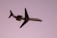 A commercial airplane, branded with Hawaiian Airline's logo and colors, is in mid-flight against a solid purple sky backdrop. The plane is well-illuminated with details visible on its fuselage and wings, suggesting it is flying during sunset or sunrise.