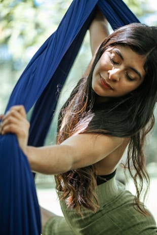 A woman with long dark hair gracefully holds onto blue aerial silk outdoors. Her eyes are closed, and she seems to be in a serene and meditative state. Sunlight filters through the leaves, creating a dappled light effect on her face and arms.