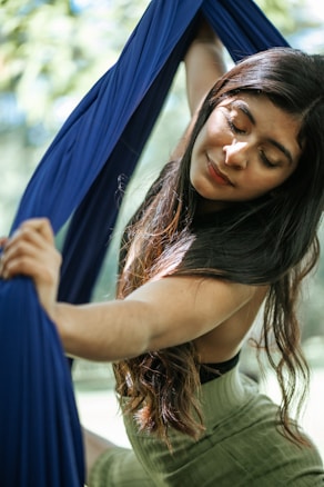 A woman with long dark hair gracefully holds onto blue aerial silk outdoors. Her eyes are closed, and she seems to be in a serene and meditative state. Sunlight filters through the leaves, creating a dappled light effect on her face and arms.