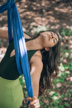 A person with long hair is gracefully holding onto blue aerial silks, surrounded by a natural outdoor setting. Sunlight filters through the trees, creating a dappled effect on the ground.