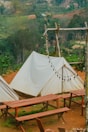 A serene outdoor setting featuring white canvas tents set up on a brown dirt ground. Wooden picnic tables and benches are arranged in front of the tents. A rustic wooden frame holds a string of unlit round bulb lights, and a lush green forest serves as the backdrop.