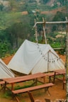 A serene outdoor setting featuring white canvas tents set up on a brown dirt ground. Wooden picnic tables and benches are arranged in front of the tents. A rustic wooden frame holds a string of unlit round bulb lights, and a lush green forest serves as the backdrop.