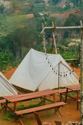A serene outdoor setting featuring white canvas tents set up on a brown dirt ground. Wooden picnic tables and benches are arranged in front of the tents. A rustic wooden frame holds a string of unlit round bulb lights, and a lush green forest serves as the backdrop.