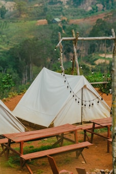 A serene outdoor setting featuring white canvas tents set up on a brown dirt ground. Wooden picnic tables and benches are arranged in front of the tents. A rustic wooden frame holds a string of unlit round bulb lights, and a lush green forest serves as the backdrop.