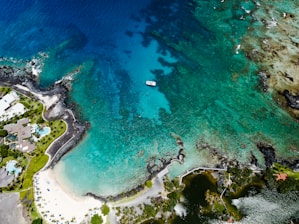 Aerial view of a coastal area with turquoise waters and a coastline filled with dark rocky edges and coral reefs. A small boat is anchored near the center of the water. On the left, an upscale housing area with modern structures and landscaped gardens is visible, along with a few swimming pools. The coastline also features sandy beaches dotted with palm trees.