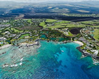Aerial view of a coastal landscape featuring vibrant blue and turquoise waters along a coastline. The image shows a sandy beach with scattered trees and various buildings, likely resorts or residential areas. Inland, lush green fields and golf courses are visible, surrounded by darker volcanic terrain.