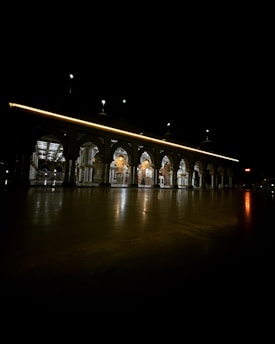 A beautifully illuminated building with a series of arches and a reflective ground surface under a dark night sky. The building features intricate architectural details and is highlighted by warm lights, creating a serene and majestic atmosphere.