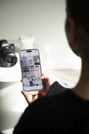 A person is holding a smartphone displaying a social media app, with various images on the screen. A camera and a disposable coffee cup are on a white surface nearby. The setting is well lit, creating a casual and focused atmosphere.