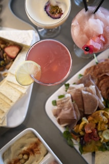An assortment of cocktails and appetizers is artfully arranged on a table. Three different cocktails are visible, each with unique garnishes. Accompanying the drinks are various appetizers, including sliced cured meats, artichoke, roasted peppers, bread, hummus, and a small selection of nuts and cheese.