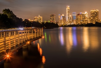 A nighttime cityscape with a brightly lit skyline reflecting on a calm body of water. A wooden boardwalk with orange lights runs along the left side, adding warmth and depth to the scene. The sky has a gentle, dim glow, indicating urban light pollution.