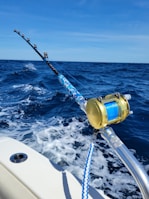 A fishing rod with a gold reel is mounted on the side of a boat, extending over the deep blue ocean. The waves create white foam as the boat moves forward under a clear blue sky.