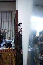 A person with a painted white face is partially hidden behind a wall, creating a sense of mystery. The setting appears to be a backstage or dressing room with colorful fabric draped over a table, various personal items, and a mirror reflecting a partially visible person in traditional attire.
