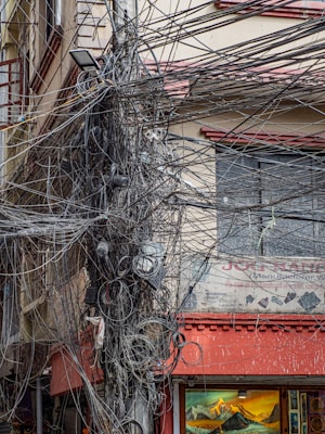 A chaotic jumble of electrical wires and cables is densely tangled around a utility pole in an urban setting. The background features a building with faded signage and windows, one of which is partially obscured by the cables. A shop on the lower level displays artwork, with one painting showcasing a mountain landscape.