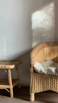 A wicker chair with a cream cushion is placed next to a wicker side table, both situated on a wooden floor. Soft natural light casts shadows on the plain white wall behind them.