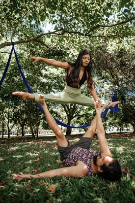 A woman performs a balanced pose on a man's feet while they both practice acro yoga in a grassy, wooded park. The ground is covered with green grass and fallen leaves, while trees with lush green foliage are visible in the background.