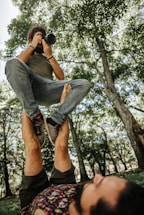 A person is balancing another person on their feet in an outdoor setting surrounded by trees. The person being balanced is holding a camera and appears to be taking a photograph. The scene takes place on a grassy area with sunlight filtering through the tree leaves.