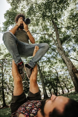 A person is balancing another person on their feet in an outdoor setting surrounded by trees. The person being balanced is holding a camera and appears to be taking a photograph. The scene takes place on a grassy area with sunlight filtering through the tree leaves.