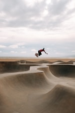 A skateboarder performs a mid-air trick above a concrete skatepark with undulating curves. The backdrop features a tranquil ocean and a sky filled with clouds.