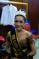A young woman wearing an ornate crown and traditional dress with intricate beadwork and embroidery sits in a room. She has a sash with gold lettering, and the background features a hanger with a white garment, along with some chairs in red and blue.