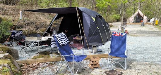 A camping setup in a wooded area includes a large dark-colored tent with an attached canopy. In front of the tent are two blue camping chairs, one draped with a black and white striped towel and the other with a flag design. A small folding table holds various camp items including a portable grill. The ground is covered with gravel, and there are trees in the background along with another tent in the distance.