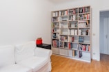 A well-organized room featuring a large white bookshelf filled with books of various sizes and colors. To the left, a white sofa is placed against the wall. A small black side table with red storage boxes sits beside the sofa. The floor is light-colored wood, and the room has a minimalistic and tidy appearance.