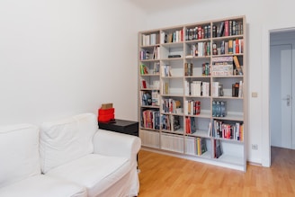 A well-organized room featuring a large white bookshelf filled with books of various sizes and colors. To the left, a white sofa is placed against the wall. A small black side table with red storage boxes sits beside the sofa. The floor is light-colored wood, and the room has a minimalistic and tidy appearance.