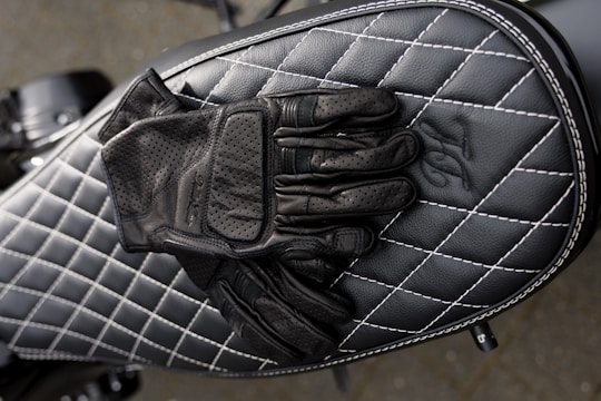 A pair of black leather gloves is placed on top of a black quilted leather motorcycle seat with white stitching. The design features diamond-shaped patterns, creating a sleek, stylish look.