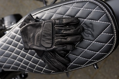 A pair of black leather gloves is placed on top of a black quilted leather motorcycle seat with white stitching. The design features diamond-shaped patterns, creating a sleek, stylish look.