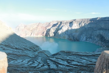 The image captures a vast volcanic crater filled with turquoise water, surrounded by rugged and textured rocky cliffs. The colors of the crater walls range from dark gray to sandy brown, contrasting with the vibrant blue of the water. Mist or smoke rises gently from the water's surface, adding an ethereal quality to the scene.