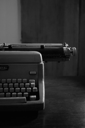 A vintage typewriter sits on a dark wooden table, partially framed in a black and white photograph. The keys are exposed, highlighting the machinery's retro design with a focus on the left side.