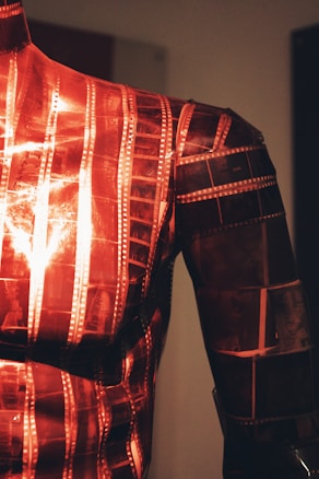 A close-up view of a mannequin or sculpture covered in overlapping strips of photographic film. The film strips are primarily red in color and reflect light in various places, creating a shiny surface.