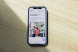A smartphone displaying a website is placed on a wooden surface. The website features text in Polish and an image of two people standing together with indoor plants in the background.