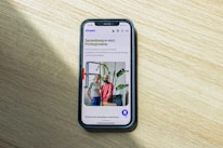A smartphone displaying a website is placed on a wooden surface. The website features text in Polish and an image of two people standing together with indoor plants in the background.