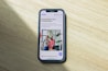A smartphone displaying a website is placed on a wooden surface. The website features text in Polish and an image of two people standing together with indoor plants in the background.