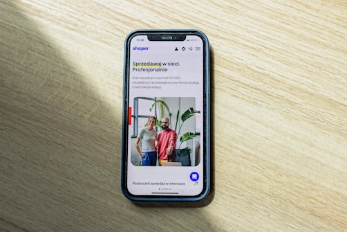 A smartphone displaying a website is placed on a wooden surface. The website features text in Polish and an image of two people standing together with indoor plants in the background.