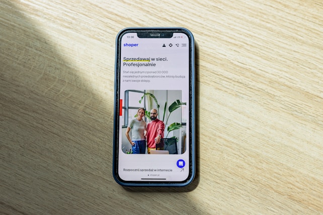 A smartphone displaying a website is placed on a wooden surface. The website features text in Polish and an image of two people standing together with indoor plants in the background.