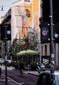 A large mural on the side of a building depicts a child reaching upwards with a paper airplane. Below, a street scene unfolds with a cyclist on an urban road. Sidewalks are lined with trees and outdoor seating under green umbrellas. Various vehicles are parked along the street, and there are buildings with visible signage.