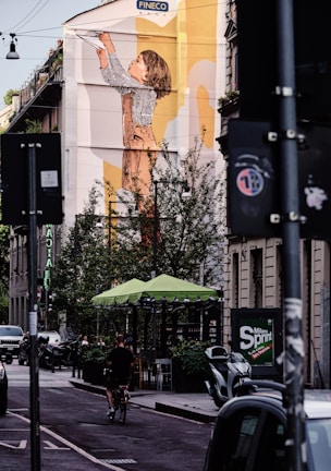 A large mural on the side of a building depicts a child reaching upwards with a paper airplane. Below, a street scene unfolds with a cyclist on an urban road. Sidewalks are lined with trees and outdoor seating under green umbrellas. Various vehicles are parked along the street, and there are buildings with visible signage.