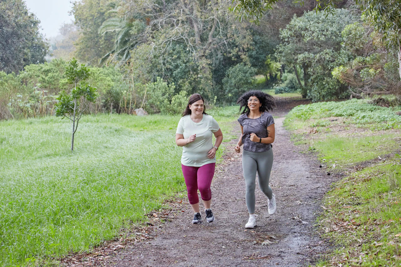 Two women smiling as they run side by side along a dirt trail through green trees