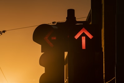 A traffic light against a warm, golden sunset background. The light displays red arrows, with one pointing up and the other to the left. The silhouette of the traffic light contrasts with the bright sky.