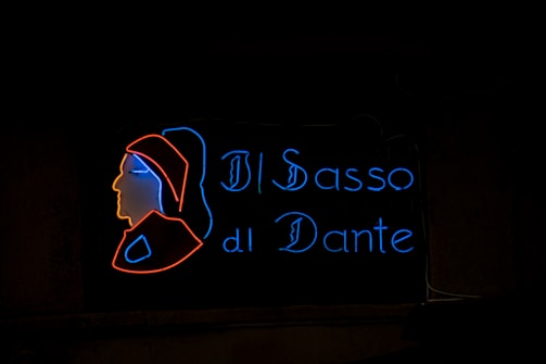 A neon sign consisting of a stylized profile silhouette outlined in red neon, accompanied by the words 'Il Sasso di Dante' in blue neon. The silhouette appears to depict a historical or artistic figure, possibly inspired by Dante Alighieri.
