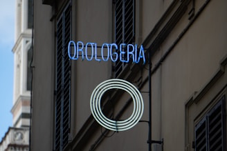 A neon sign reading 'Orologeria' with a circular pattern is mounted on the exterior of a building next to shuttered windows. The structure in the background appears to be part of a historical tower with ornate architectural details.