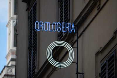 A neon sign reading 'Orologeria' with a circular pattern is mounted on the exterior of a building next to shuttered windows. The structure in the background appears to be part of a historical tower with ornate architectural details.