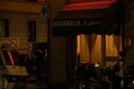 A dimly lit street scene featuring the entrance to a pizzeria named Pizzeria S. Lucia. The awning is dark green, and soft yellow light emanates from inside, casting a warm glow. Several parked motor scooters are visible along with a small vehicle, surrounded by urban architecture.