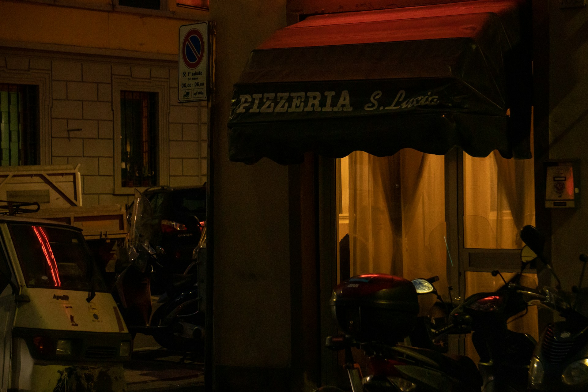 A dimly lit street scene featuring the entrance to a pizzeria named Pizzeria S. Lucia. The awning is dark green, and soft yellow light emanates from inside, casting a warm glow. Several parked motor scooters are visible along with a small vehicle, surrounded by urban architecture.