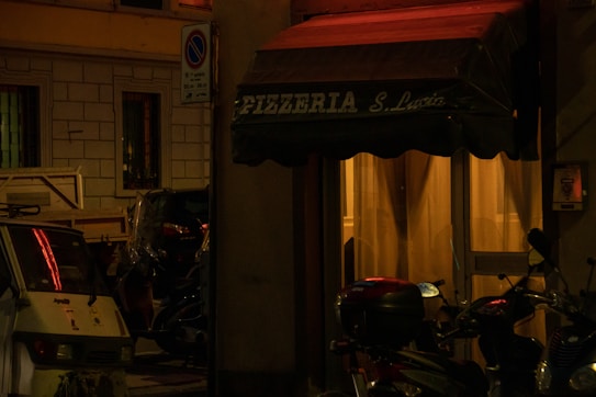 A dimly lit street scene featuring the entrance to a pizzeria named Pizzeria S. Lucia. The awning is dark green, and soft yellow light emanates from inside, casting a warm glow. Several parked motor scooters are visible along with a small vehicle, surrounded by urban architecture.