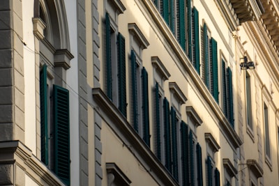 A series of windows with dark green shutters are set in a beige or cream-colored building wall with decorative molding. The building features classic architectural elements, including arched and rectangular window frames, and protruding ledges.