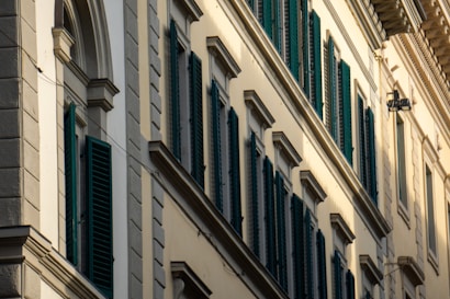 A series of windows with dark green shutters are set in a beige or cream-colored building wall with decorative molding. The building features classic architectural elements, including arched and rectangular window frames, and protruding ledges.